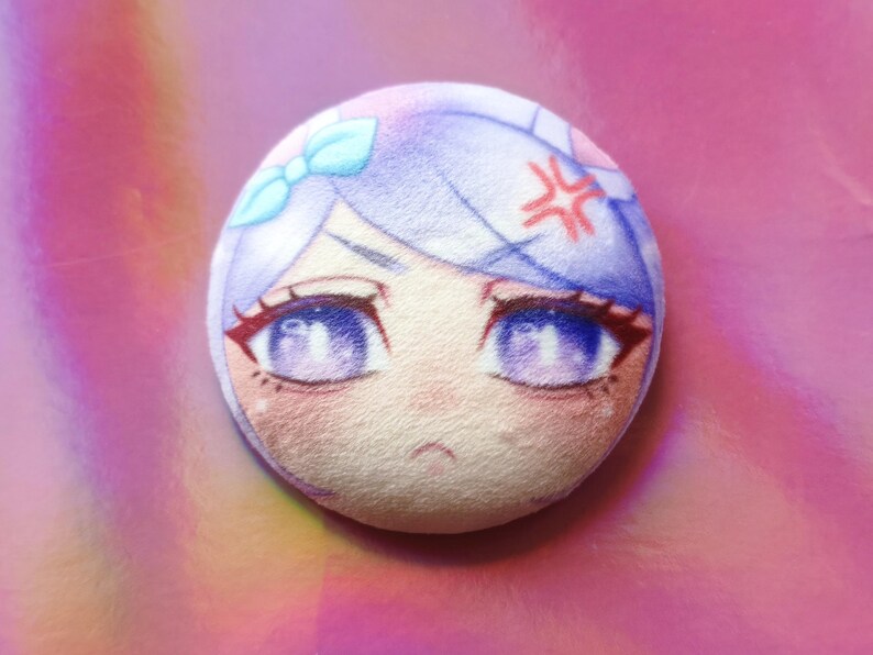 May include: A circular badge displaying an anime-style character's face with purple eyes, light blue hair, and a red angry symbol. The badge has a soft, textured appearance and a light pink background.