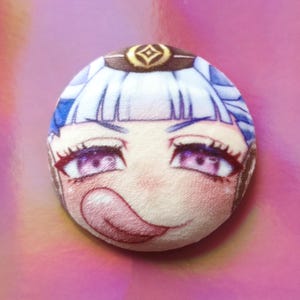 May include: A round plushie with an anime-style face, light blue hair, purple eyes, and a protruding tongue. It has a brown and gold headpiece, set against a pink background. The plushie is a cute, cartoon-like design.