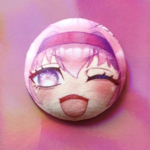 May include: A circular plush toy with a pink anime-style face, winking eye, rosy cheeks, and a smiling expression. The toy has pink hair and a headband. The background is a gradient of pink and purple.