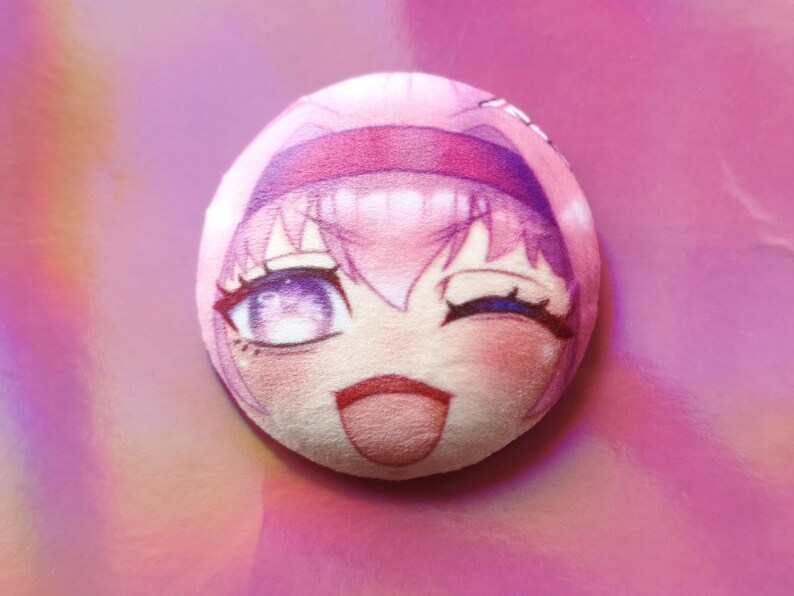 May include: A circular plush toy with a pink anime-style face, winking eye, rosy cheeks, and a smiling expression. The toy has pink hair and a headband. The background is a gradient of pink and purple.