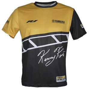 May include: A yellow and black t-shirt with a white Yamaha logo and the text "Factory Racing". The shirt also features a white "R1" logo, a white signature that says "Kenny Roberts", and a black and white striped design.