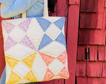May include: A square quilted tote bag featuring a patchwork design of cream-colored fabric interspersed with floral-patterned triangles in shades of lavender, blue, yellow, and peach. The bag has two fabric shoulder straps and a stitched texture.