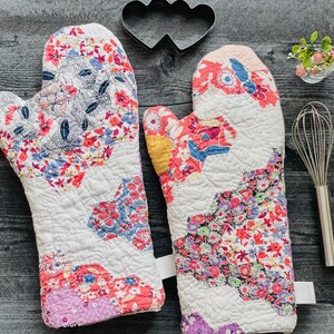 May include: A pair of quilted oven mitts featuring a patchwork design with floral and abstract patterns in shades of pink, red, blue, yellow, and white. The mitts are accompanied by a black metal heart-shaped cookie cutter and a metal whisk.