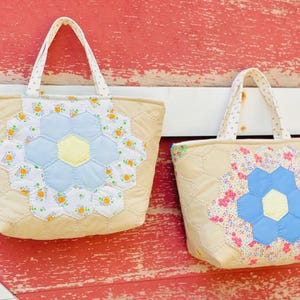 May include: Two beige quilted tote bags with floral appliqués. The bags feature a large flower design in the center, with blue and yellow petals. The handles are white with a polka dot pattern.