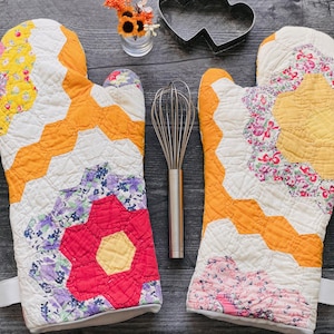 May include: A pair of quilted oven mitts featuring a colorful patchwork design of hexagons in yellow, orange, red, and purple floral patterns. A stainless steel wire whisk and a heart-shaped metal cookie cutter are positioned between the mitts.