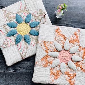May include: Two square quilted zipper pouches featuring appliquéd flower designs. One pouch has a yellow center with teal petals on a beige star pattern, while the other has a pink center with white petals on an orange star pattern.