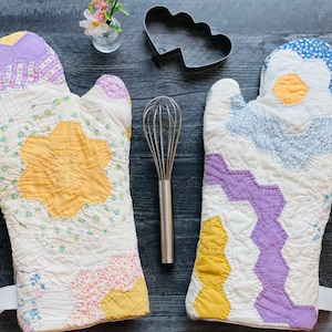 May include: Two quilted oven mitts featuring a patchwork design of hexagonal floral patterns in yellow, lavender, blue, and white. The mitts are accompanied by a metal whisk, a heart-shaped cookie cutter, and a small glass vase with flowers.