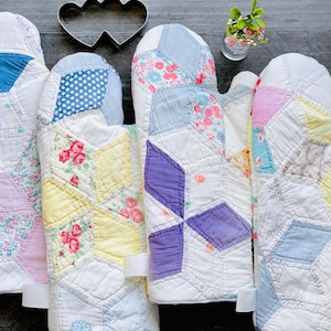 May include: Four quilted oven mitts featuring a patchwork design of various fabrics including white, blue, yellow, pink, and purple patterns with floral and polka dot prints. Each mitt has a white fabric loop attached to the side for hanging.