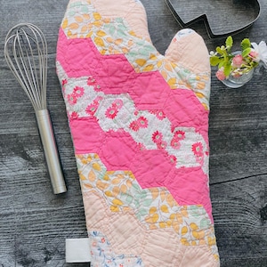 May include: A quilted oven mitt featuring a patchwork of hexagonal fabric pieces in shades of pink, peach, yellow, and white with floral and leaf patterns. The mitt has a white fabric loop at the base and visible stitching throughout the quilted surface.