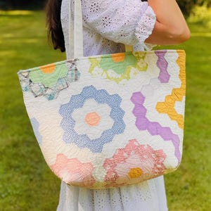 May include: A tote bag constructed from quilted fabric featuring a patchwork design of hexagonal floral patterns in pastel shades of blue, pink, yellow, green, and purple on a white background, equipped with two white fabric shoulder straps.