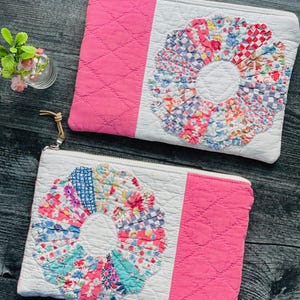 May include: Two rectangular zippered pouches featuring a white quilted fabric base with a pink quilted side panel. Each pouch displays a circular floral applique made from various colorful patterned fabric scraps and a tan leather zipper pull.