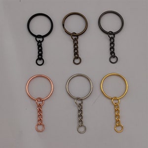 Könnte beinhalten: Sechs Schlüsselringe mit befestigten Ketten in verschiedenen Farben: Schwarz, Silber, Gold, Roségold. Die Schlüsselringe sind alle gleich groß und gleich geformt, und die Ketten sind alle gleich lang.