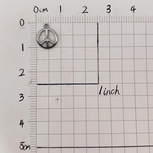 May include: A silver peace sign charm, measuring approximately 1 inch in diameter, is shown on a gridded surface.