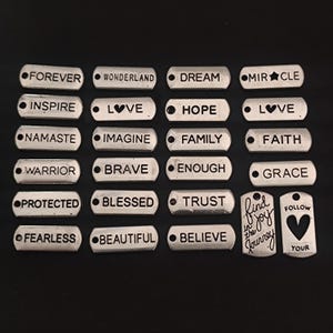 May include: A set of 18 silver-toned metal tags with various inspirational words and phrases, including "Forever", "Love", "Hope", "Family", "Faith", "Warrior", "Brave", "Enough", "Grace", "Protected", "Blessed", "Trust", "Fearless", "Beautiful", "Believe", "Find the Joy", and "Follow Your Heart".