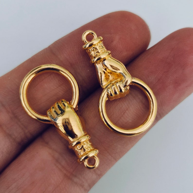 K&ouml;nnte beinhalten: Zwei goldfarbene, handf&ouml;rmige Ringz&uuml;ge aus Metall mit einer runden Schlaufe. Die Hand h&auml;lt die Schlaufe.