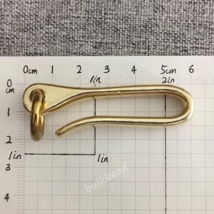 May include: A gold-colored metal hook with a rounded loop at one end and a curved hook at the other. The hook is approximately 5 cm long and is measured against a ruler.