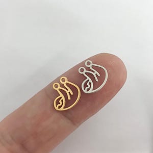 May include: Two metal sloth charms, one gold and one silver, on a finger. The charms are simple line drawings of a sloth.