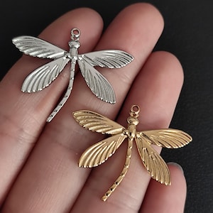 Op de afbeelding: Twee libellenbedels, één zilverkleurig en één goudkleurig, worden getoond. De bedels hebben gedetailleerde vleugelontwerpen en een lus aan de bovenkant om aan sieraden of andere knutselwerken te bevestigen. De libellenbedels worden in een hand gehouden.
