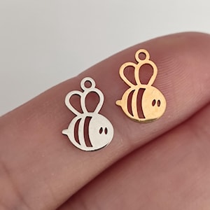 May include: Two small bee-shaped metal charms, one in a silver tone and one in a gold tone. Each charm features a cutout design with a rounded body, two wings, and a small circular loop at the top for attachment, showcasing a flat, thin profile.