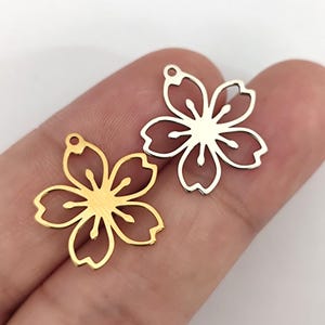 Peut inclure: Deux breloques en forme de fleur de cerisier, l'une dorée et l'autre argentée. Les breloques ont un design délicat et complexe et sont parfaites pour la fabrication de bijoux.