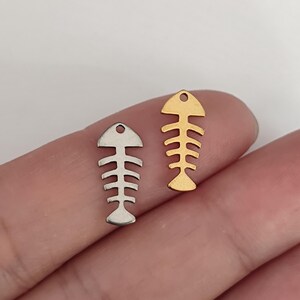 May include: Two small metal fish skeleton charms, one in a silver tone and one in a gold tone. Each charm features a flat, stylised design with a small circular hole at the top for attachment, showcasing a detailed spine and rib structure.