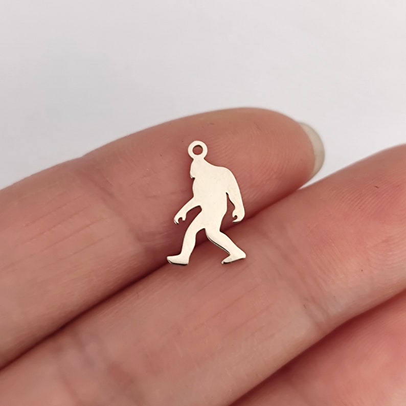 May include: Silver charm in the shape of a Bigfoot walking. The charm has a hole at the top for attaching to a necklace or bracelet.