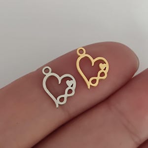 Könnte beinhalten: Zwei herzförmige Anhänger, einer in Silberoptik und einer in Goldoptik. Jeder Anhänger besteht aus einer offenen Herzform mit einem kleinen ausgefüllten Herz und einem Unendlichkeitssymbol an der unteren rechten Seite sowie einer kleinen Öse oben.