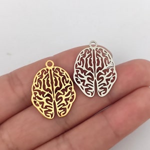 May include: Two metal brain charms, one gold and one silver, with a detailed outline of a human brain. The charms have a loop at the top for attaching to jewellery.