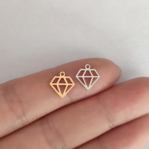 May include: Two diamond-shaped charms, one gold-colored and one silver-colored, are displayed. The charms have a geometric design with a small loop at the top for attachment. The background is a neutral, light color.