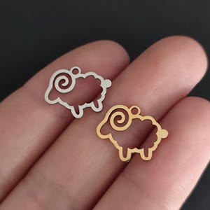 Op de afbeelding: Twee schaapvormige bedels. Eén is zilverkleurig, de andere goudkleurig. Beide hebben een lus om op te hangen en een uitgesneden ontwerp. De bedels worden in een hand gehouden tegen een donkere achtergrond.