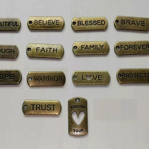 May include: A set of 13 gold-toned metal tags with various words and phrases, including "Beautiful", "Believe", "Blessed", "Brave", "Enough", "Faith", "Family", "Forever", "Hope", "Warrior", "Love", "Protected", "Trust", and "Follow Your Heart".