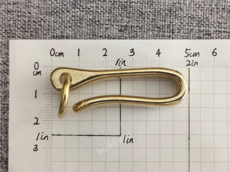 May include: A gold-colored metal hook with a rounded opening. The hook is about 5 cm long and is measured against a ruler.