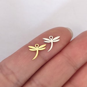 Pode incluir: Dois pingentes de libélula, um dourado e outro prateado, são mostrados em um dedo. Os pingentes são pequenos e delicados, com um design simples.