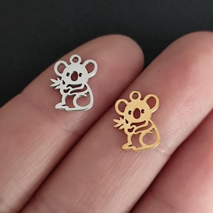 May include: Two koala bear charms, one silver and one gold, are displayed. The charms are cut-out designs, each holding a sprig of leaves. The koalas have a small loop at the top for attaching to jewelry.