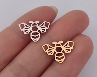 May include: Two geometric bee charms, one gold and one silver, are shown on a hand. The bee charms are made of metal and have a detailed design.
