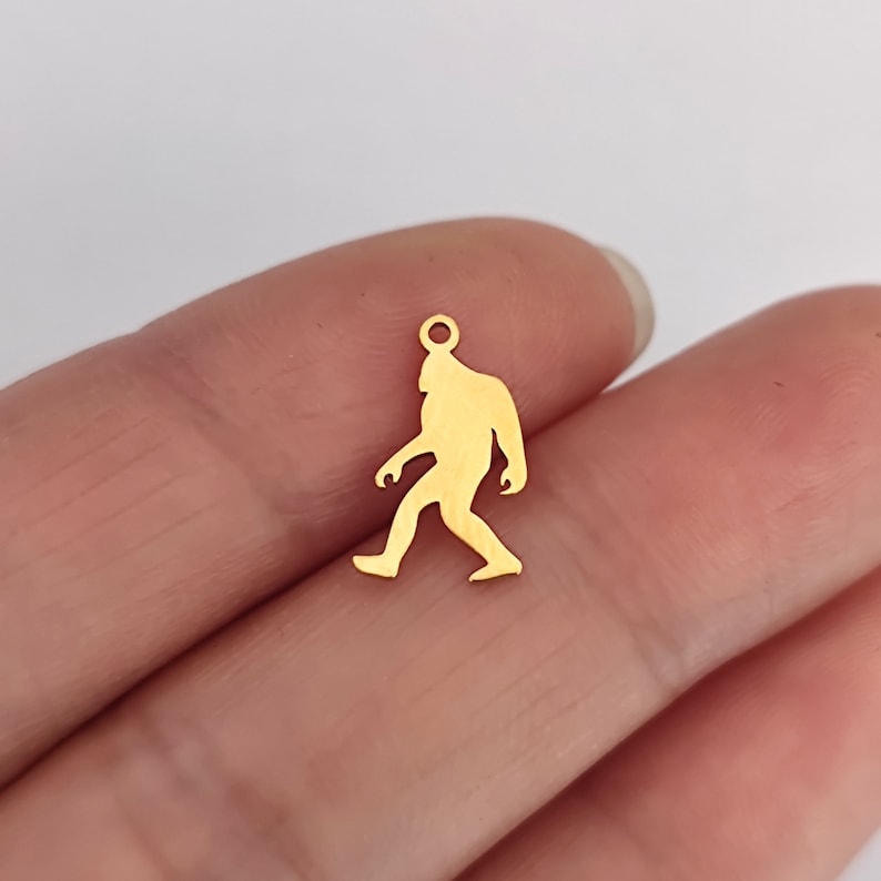 May include: Gold-colored metal charm shaped like a Bigfoot walking. The charm has a loop at the top for attaching to a necklace or bracelet.
