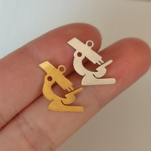 Puede incluir: Dos pequeños colgantes de microscopio. Uno es de color dorado, el otro plateado. Ambos colgantes tienen un pequeño lazo en la parte superior para sujetarlos a joyas. El diseño del microscopio es un contorno simplificado.