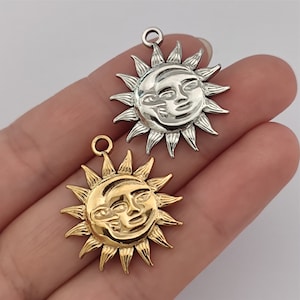 Pode incluir: Dois pingentes de sol, um dourado e outro prateado, com um design de rosto. Os pingentes estão sobre um fundo branco.