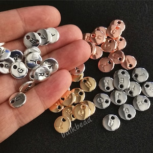 May include: Assortment of silver, gold, and rose gold metal letter and number charms with two holes. The charms are round and have a smooth, shiny finish.