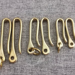 May include: Seven brass keychains with a hook shape. The keychains are all the same size and shape, but they have different finishes. Some are polished, while others are brushed.