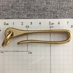 May include: A brass hook with a rounded loop at one end and a curved hook at the other. The hook is approximately 2 inches long and is measured against a ruler.