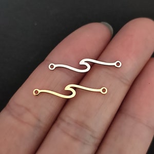 May include: Two wave-shaped metal charms with a small loop at each end. The charm on top is silver, and the charm on the bottom is gold.