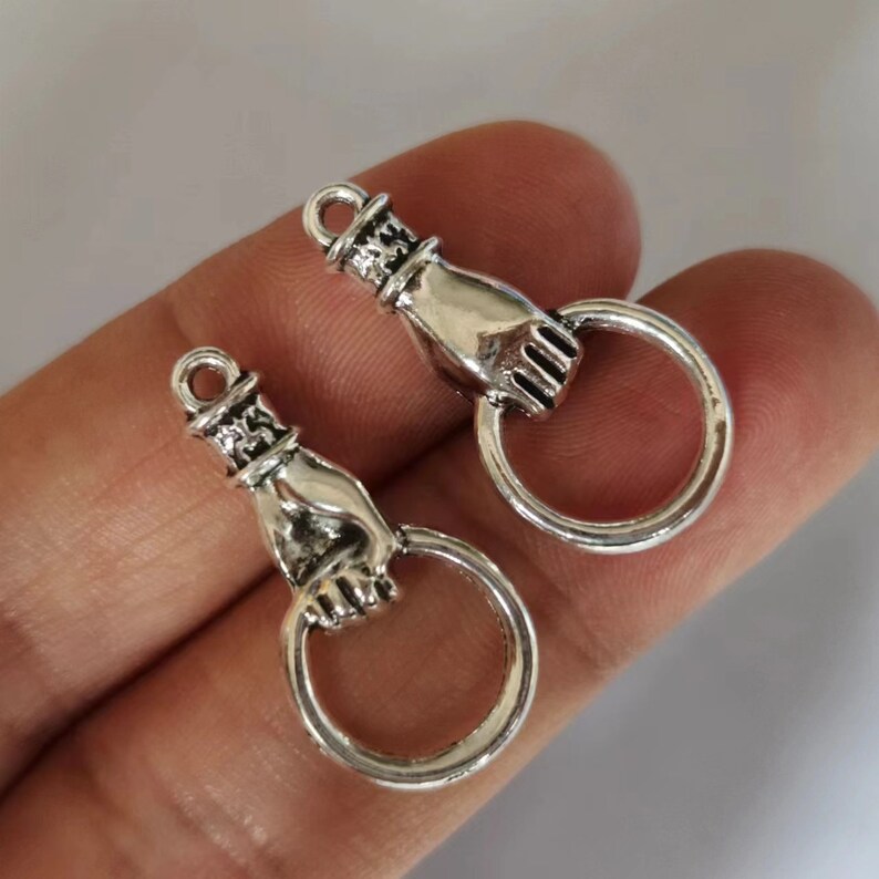 K&ouml;nnte beinhalten: Zwei silberfarbene handf&ouml;rmige Anh&auml;nger mit einem Ring an jeder Hand. Die Anh&auml;nger sind so gestaltet, dass sie wie eine Hand aussehen, die einen Ring h&auml;lt.
