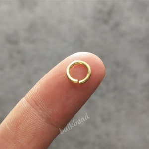 Puede incluir: Un anillo de salto dorado, un peque&ntilde;o anillo de metal utilizado para conectar componentes de joyer&iacute;a, se sostiene entre un dedo y el pulgar.