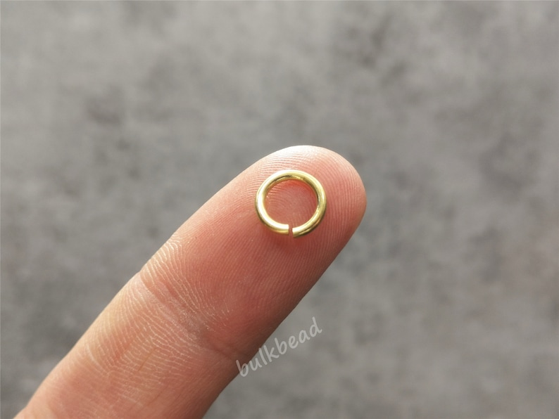 Puede incluir: Un anillo de salto dorado, un peque&ntilde;o anillo de metal utilizado para conectar componentes de joyer&iacute;a, se sostiene entre un dedo y el pulgar.