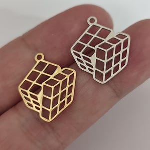 Könnte beinhalten: Zwei Würfelanhänger aus Metall, einer goldfarben und einer silberfarben, werden gezeigt. Jeder Anhänger hat ein ausgeschnittenes Design, das einem Rubik's Cube ähnelt. Die Anhänger haben oben eine kleine Schlaufe zum Befestigen an einer Kette oder einem anderen Schmuckbestandteil.