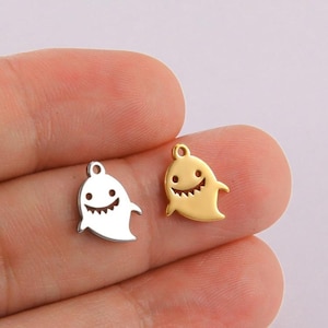May include: Two metal charms in the shape of a ghost with a smiling face. One charm is gold and the other is silver.
