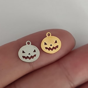 May include: Two small metal charms shaped like circular jack-o'-lanterns with cut-out facial features. One charm is silver-toned and the other is gold-toned. Each piece features a small circular loop at the top for attachment to jewelry or crafts.