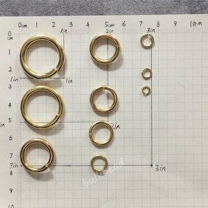 Puede incluir: Surtido de anillas de salto de metal de color dorado en varios tama&ntilde;os, medidos en pulgadas y cent&iacute;metros, sobre un fondo cuadriculado blanco.