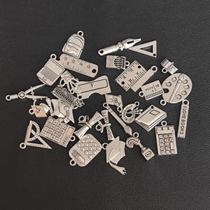 Könnte beinhalten: Eine Sammlung silberfarbener Metall-Anhänger mit Schulmotiven, darunter eine Abschluss-Kappe, ein Lineal, eine Farbpalette, ein Taschenrechner, ein Buch und ein Bleistift. Die Anhänger sind perfekt, um Schmuck oder Bastelarbeiten eine persönliche Note zu verleihen.